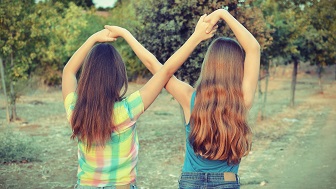 Zwei Mädchen mit langen Haaren haben die Hände gemeinsam über ihren Köpfen gekäuzt. Sie stehen draußen in einem Garten. 